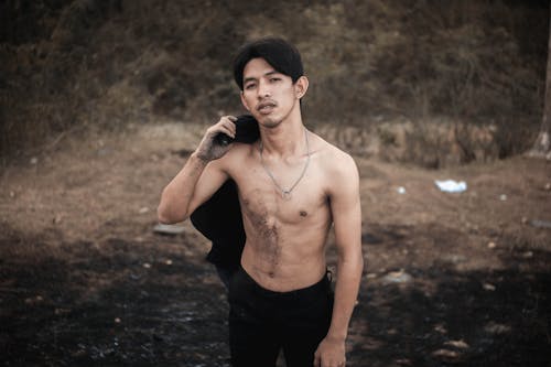 Self assured young Asian guy with dirty hands and naked torso standing in forest against leafless trees and looking at camera