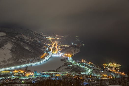 From above of glowing night town with snowy hills in heavy snowfall at night