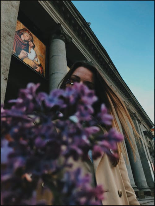 From below of anonymous woman covering face with lilacs standing on against old building in city