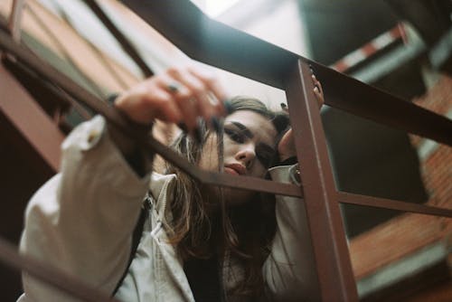 A Woman in Beige Coat Sitting Near Metal Railings while Looking at the Camera