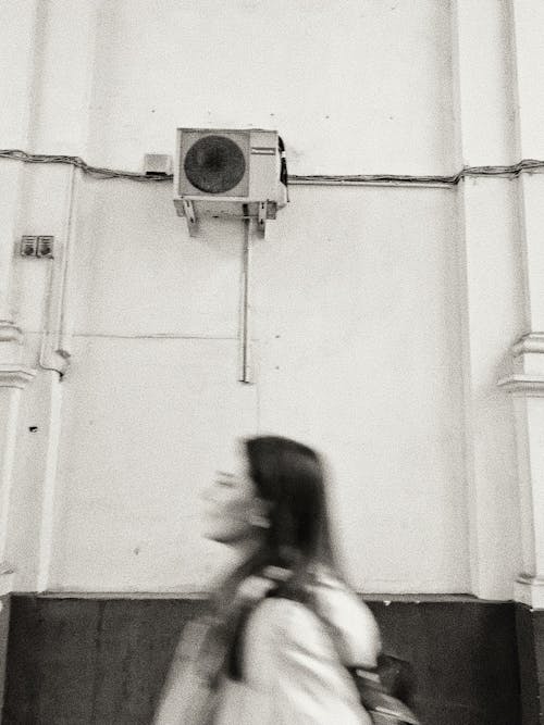 Black and white side view of young female walking near shabby wall of building with air conditioning