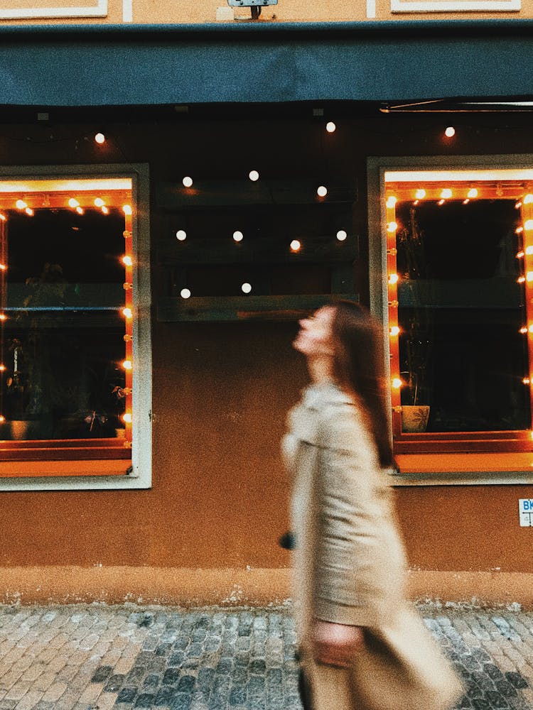 Young Woman In Cloak Walking On Sidewalk Along Luminous Building