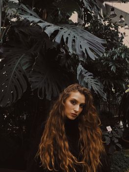 一位年轻女子留着长长的卷发，站在郁郁葱葱的室内花园中，散发出宁静而沉思的氛围
