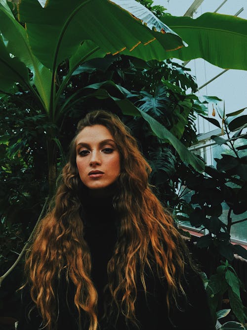 Confident young female with long curly hair and stylish makeup standing against various green plants in greenhouse