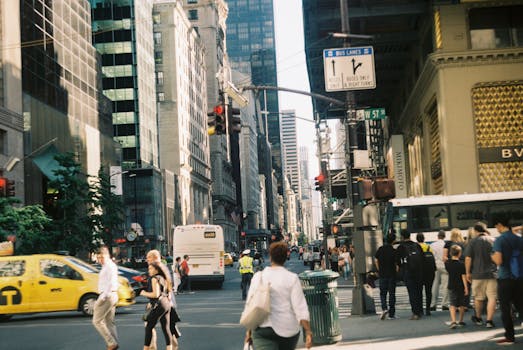Busy city street with pedestrians, cabs, buses, and tall buildings on a sunny day.