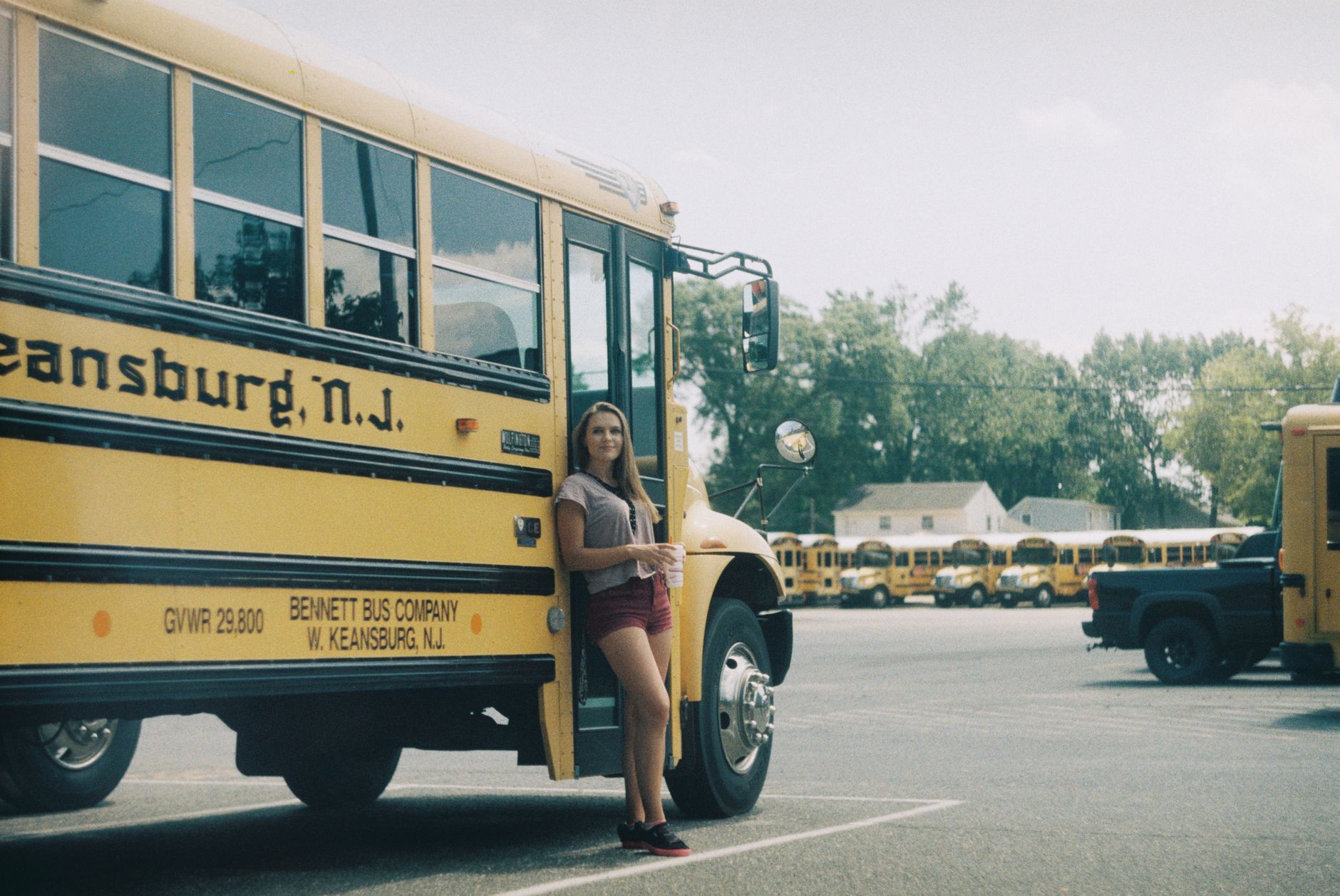 Full body of smiling female in summer clothes leaning on yellow bus on parking lot
