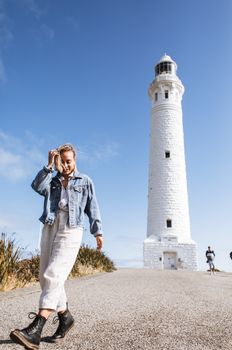 一位穿着休闲服装的女性旅行者整理着头发，走在高大的白砖灯塔旁的道路上，头顶是阳光照耀下充满活力的蓝天