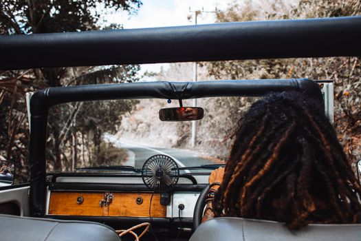 Back view of unrecognizable traveler with stylish dreadlocks spinning steering wheel while driving car and reflecting in small mirror
