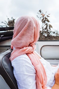 Side view of faceless female in light pink headscarf looking at dark green foliage while driving car in daytime on blurred background