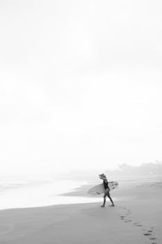 A serene black and white photograph of a woman surfer walking along a tranquil beach.