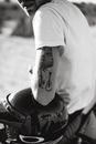 Black and white crop biker in white shirt with tattoo on arm putting hand on glossy helmet in sunlight on blurred background