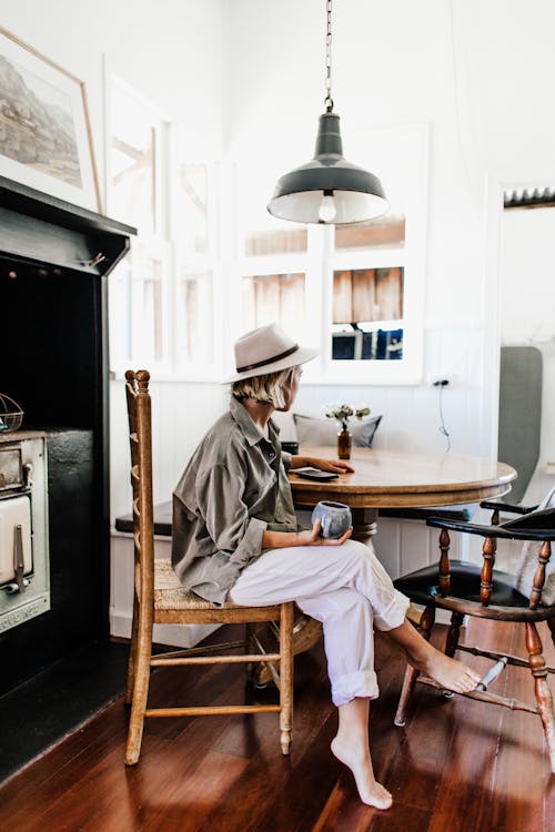 Full body side view of young unrecognizable barefoot female in casual clothing and hat drinking coffee while lounging at round wooden table in bright cozy kitchen