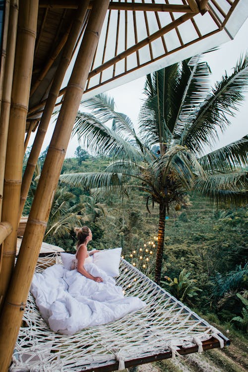 Side view of faceless young slender female with bunch on head looking away while chilling on unusual bed under blanket in tropical country