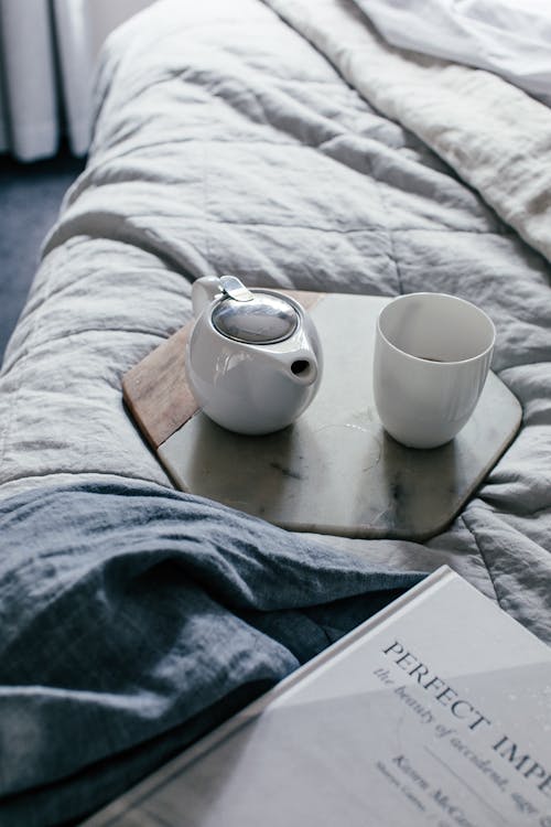 From above of white cup and teapot on tray next to book on blanket in bedroom in daytime