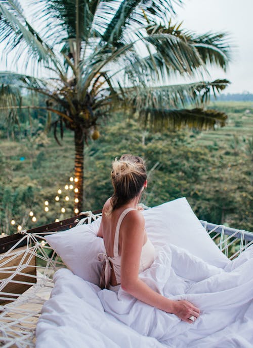 Back view of unrecognizable young slender female tourist chilling on white fabric mesh and looking away in tropical country