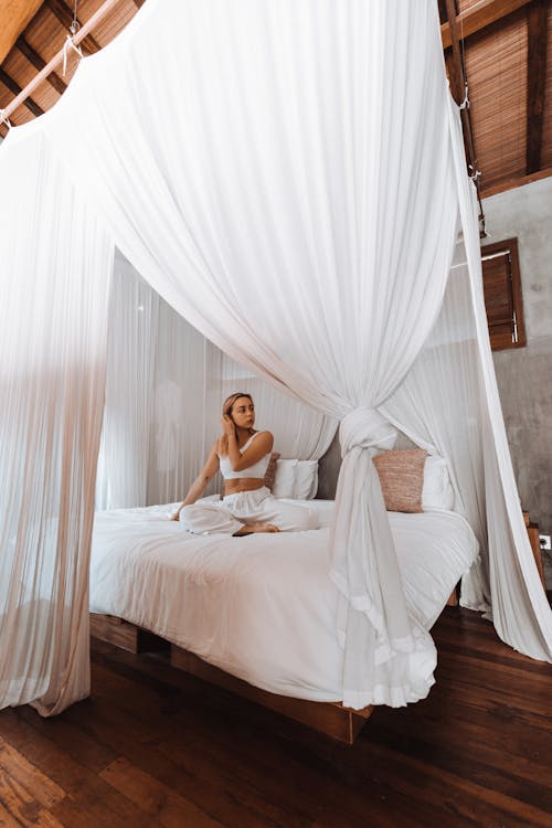 Free Full body of charming female in sleepwear sitting on comfortable canopy bed in cozy room Stock Photo