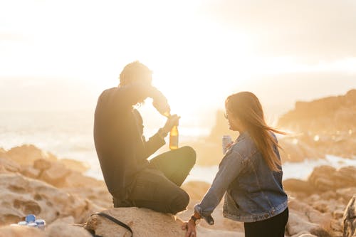 Side view of faceless couple in casual clothes drinking beer while resting on stones near ocean at sundown
