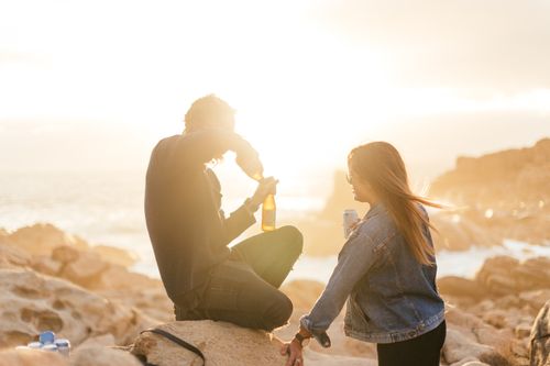 Free Side view of faceless couple in casual clothes drinking beer while resting on stones near ocean at sundown Stock Photo