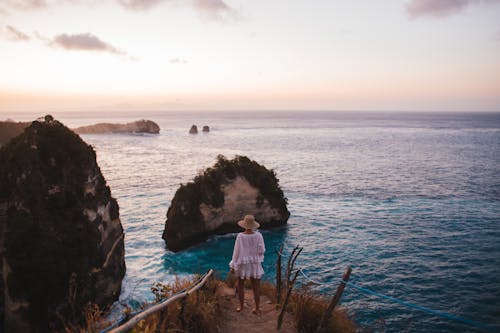 Back view of faceless female in straw hat and summer clothes standing on shore near rock formations and endless ocean