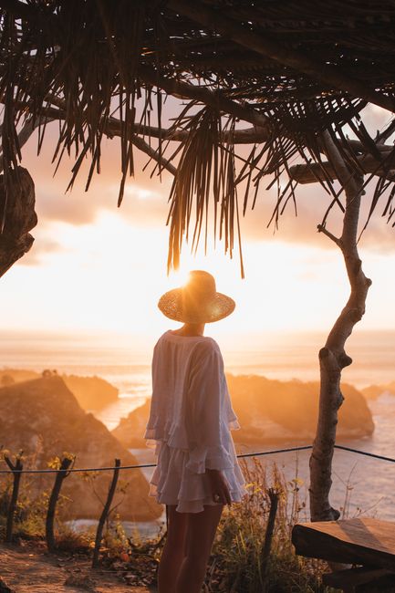 Back view of faceless female in straw hat standing on mountain over cliffs and ocean at sundown