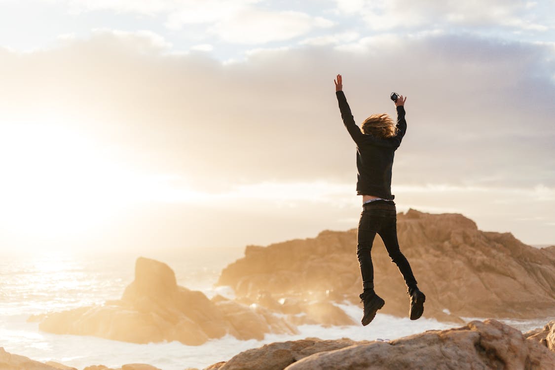 Free Full body of anonymous male in casual clothes jumping on coast near ocean while spending time at sunset Stock Photo