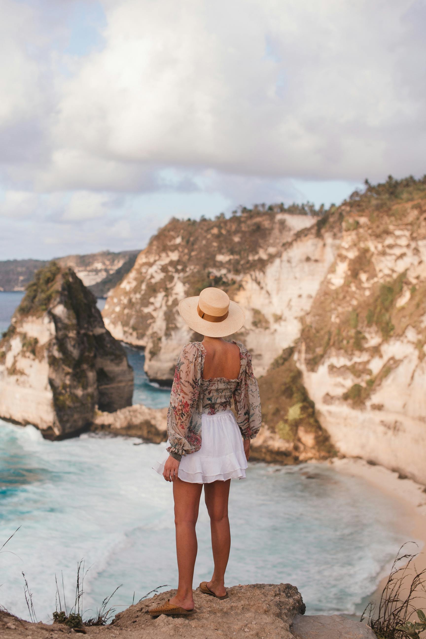 Full body of stylish unrecognizable female in straw hat and summer clothes standing on rocky mountain over foaming ocean
