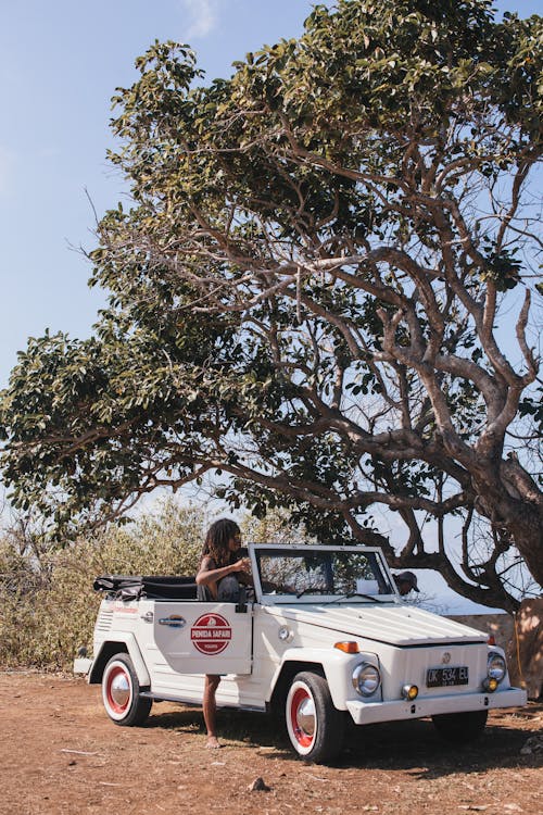 Full body of faceless African American person standing near convertible car under tree with lush tropical vegetation