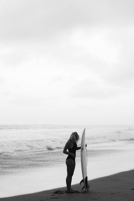 Captivating black and white photo of a woman holding a surfboard on a beach with waves in the background.