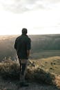 一位无名男游客背对镜头，手插口袋，在多云的天空下欣赏着草甸附近的灌木丛