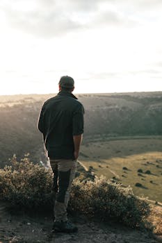 一位无名男游客背对镜头，手插口袋，在多云的天空下欣赏着草甸附近的灌木丛
