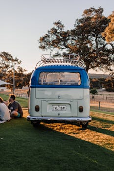 Rear bumper of vintage van on green lawn near crop anonymous friends and trees in soft sunlight