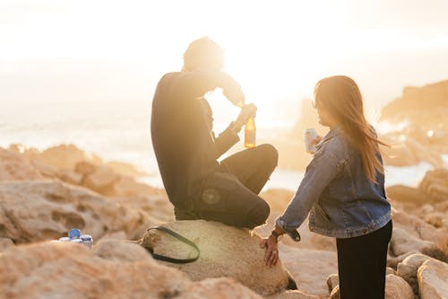Side view of anonymous couple drinking alcohol beverages while spending time on stony shore in sunlight with backlit on blurred background