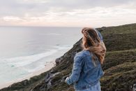 Back view of unrecognizable female with flying hair standing near sea on edge of rocky coast with hand on head and looking into distance