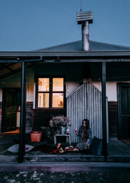 Free Relaxed female with glass of wine sitting on wooden porch of residential cottage near table with flowers while enjoying dinner outside house in evening time Stock Photo