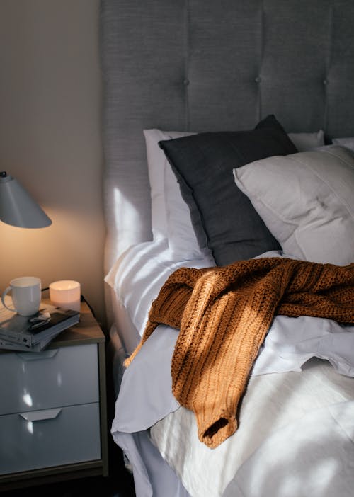 Cozy bed with pillows and knitted sweater near bedside table with mug on books in hotel room