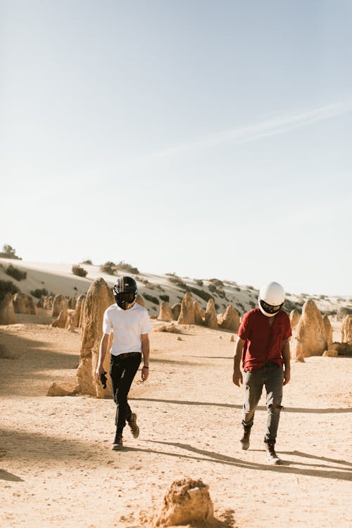 Anonymous male bikers in casual apparel strolling on dry sandy land with rocky formations under cloudy sky