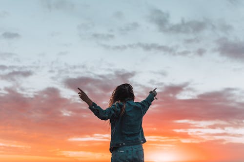 Free Back view of anonymous female in denim apparel showing colorful cloudy sky with fingers while contemplating sundown Stock Photo