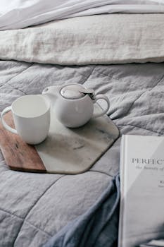 From above of mug near small teapot on marble and wooden tray on soft bed in hotel room