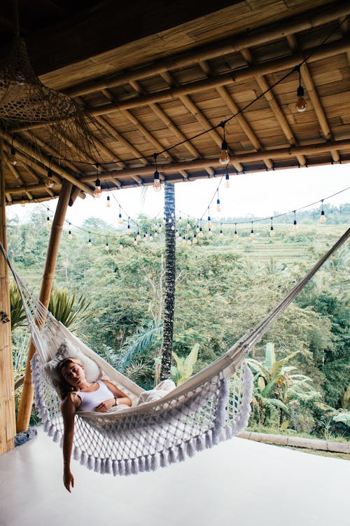 Young tired female tourist lying in hammock with fringes on bamboo veranda against exotic plants while looking at camera
