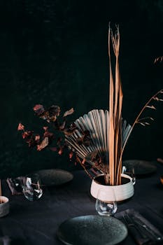 Table setting with dishware and cutlery near vase with decorative dried palm leaves and flowers
