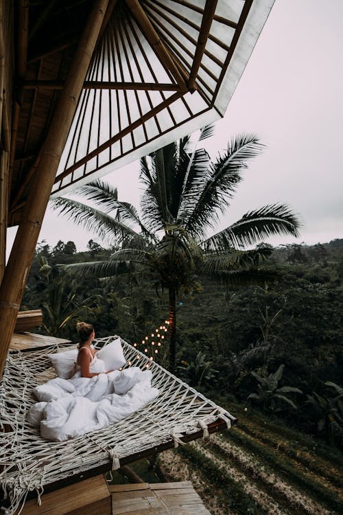 From above side view of anonymous female traveler in white blanket relaxing in hammock and admiring view of lush dense woods