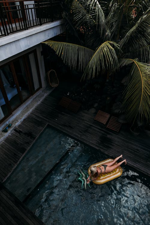 From above back view of anonymous female in swimwear lying on air mattress in pool of villa at tropical resort