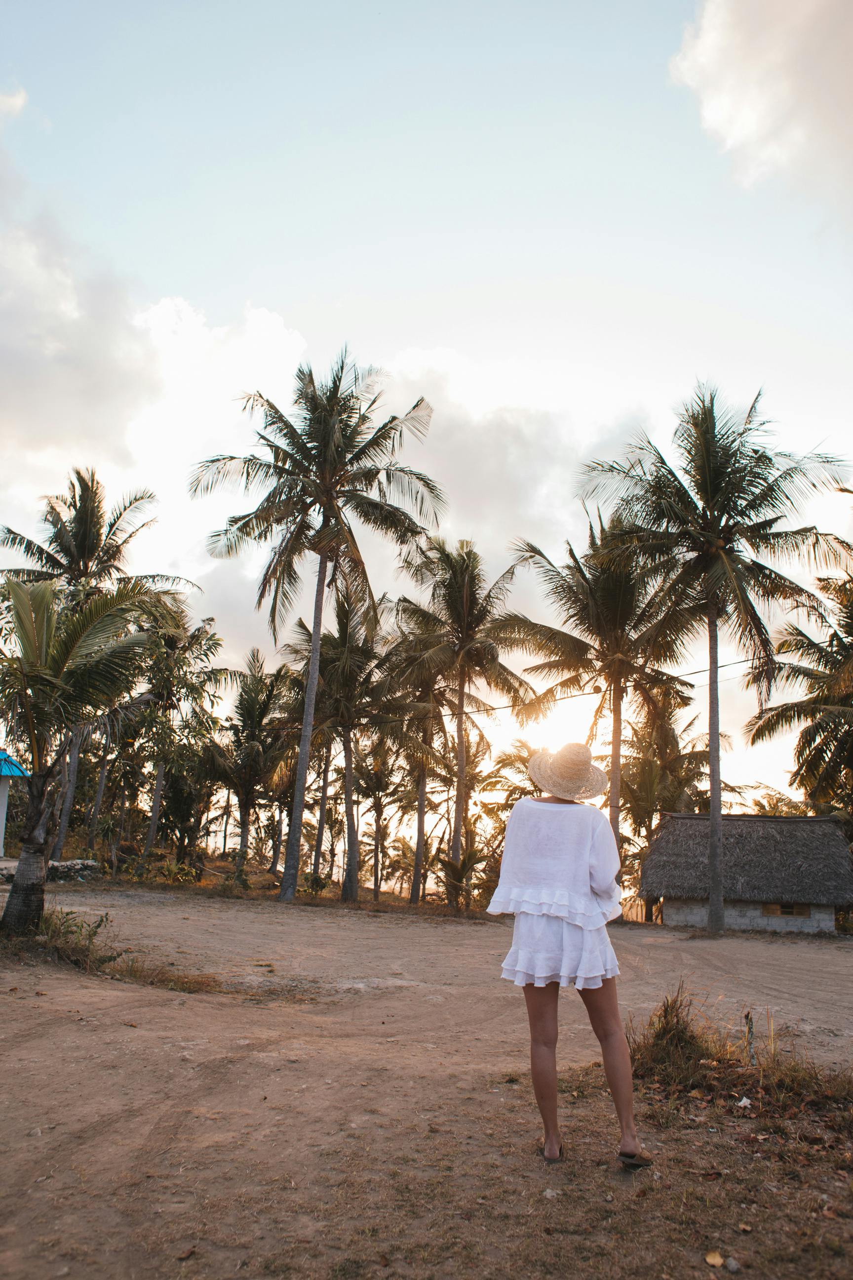 日落时分，一位匿名女游客站在空旷的沙质海岸上，身后是热带棕榈树