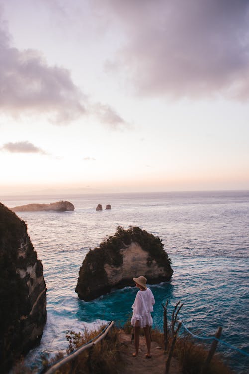 High angle back view of faceless female traveler wearing straw hat standing near rippling sea at sundown