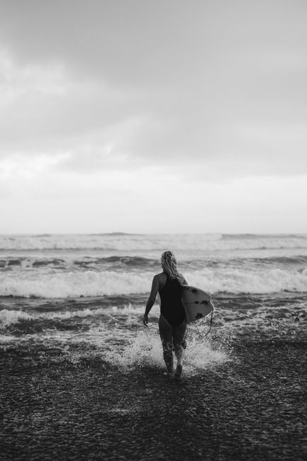 Black and white full length of female tourist with surfboard walking in splashing foamy waves