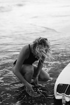 Black and white of female surfer in swimwear sitting in foamy water of sea while fastening leash on leg