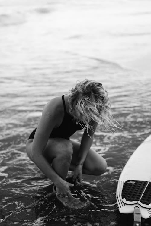 Black and white of female surfer in swimwear sitting in foamy water of sea while fastening leash on leg
