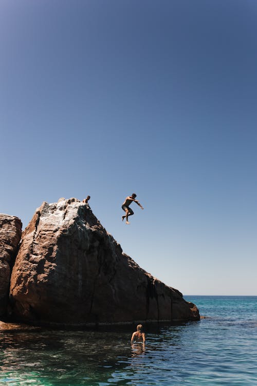 Unrecognizable man jumping off cliff into clear blue water on summer sunny day