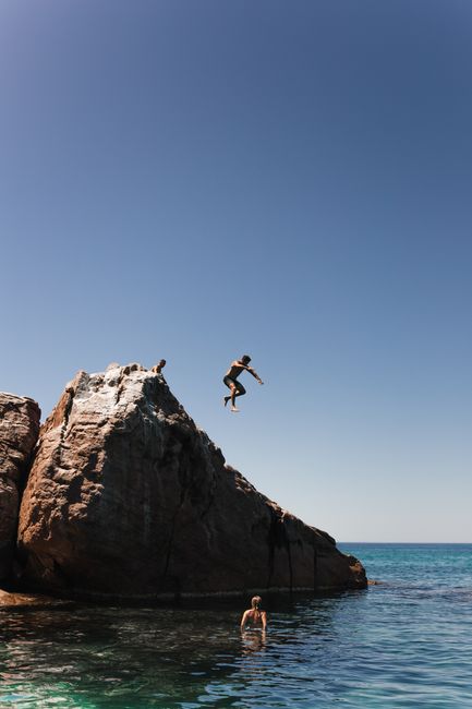 Unrecognizable female tourist standing in rippling blue water and watching how male partner cliff jumping into clear blue ocean on summer day