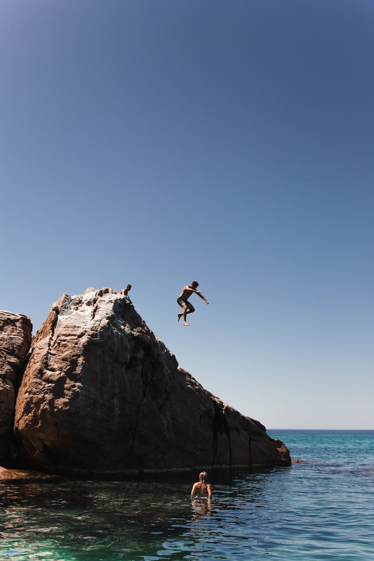 Unrecognizable Man Jumping Off Cliff Into Clear Blue Water On Summer Sunny Day